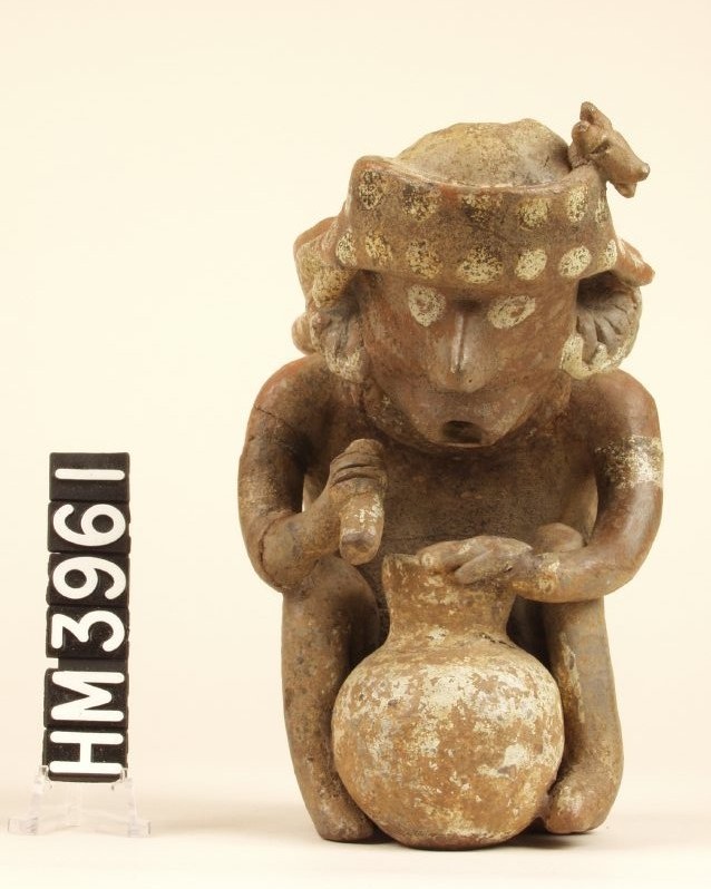 Small ceramic figure with headdress seen from the front sitting with jar between knees. Mouth pursed. Implement in right hand.