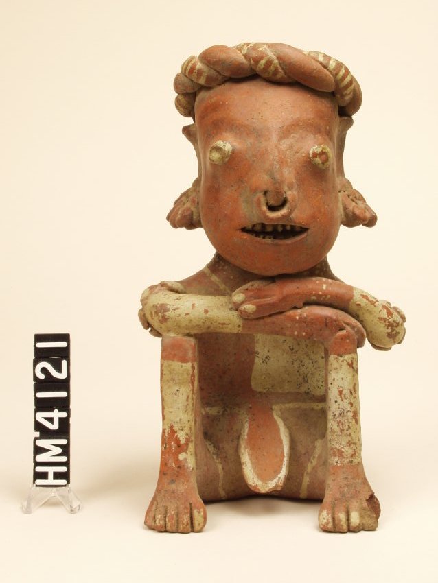 Ceramic figure seated with both legs bent and drawn up to chest; feet flat on the ground. Arms are crossed and rest on knees.