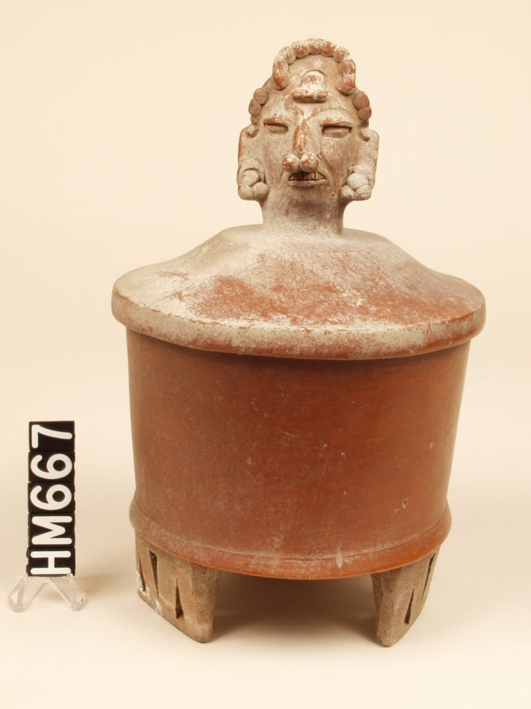 Ceramic covered urn on wide rectangular feet. Cover has decorative head modeled center top.
