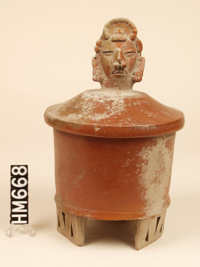 Ceramic covered urn on wide rectangular feet. Cover has decorative head modeled center top.