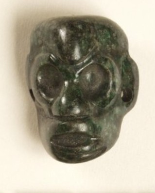 Small greenstone bead in the shape of a face, mouth slightly open, eyes possibly frowning.