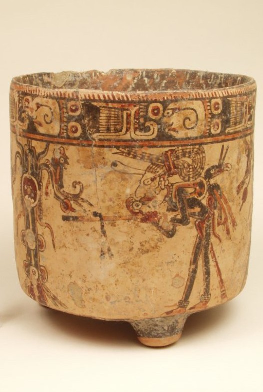 Ceramic vase, taller than wide with vertical sides and one of three small feet visible. Decorative band just below rim and intricate figures painted below.