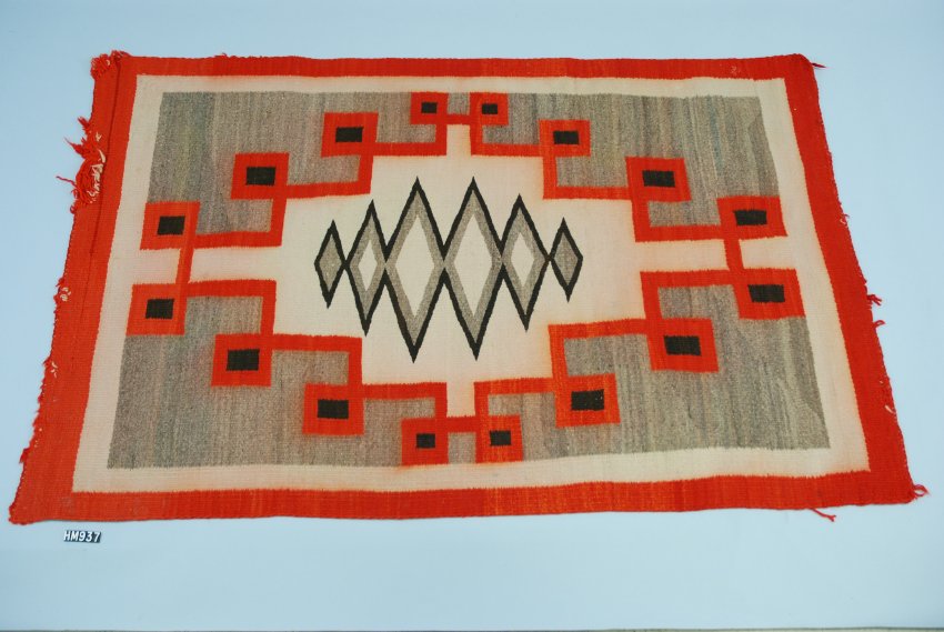 A large woven rug in red, gray, black, and cream. An array of diamonds in the center with framed by an angular line creating squares and a stepped outline.