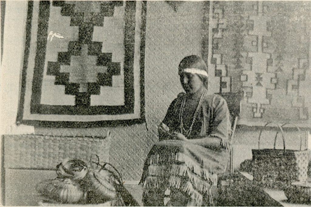 Black and white image of a woman sitting in a room. She wears a dress with strips of fringe and a headband. She is surrounded by baskets and woven textiles hang on the wall behind her.