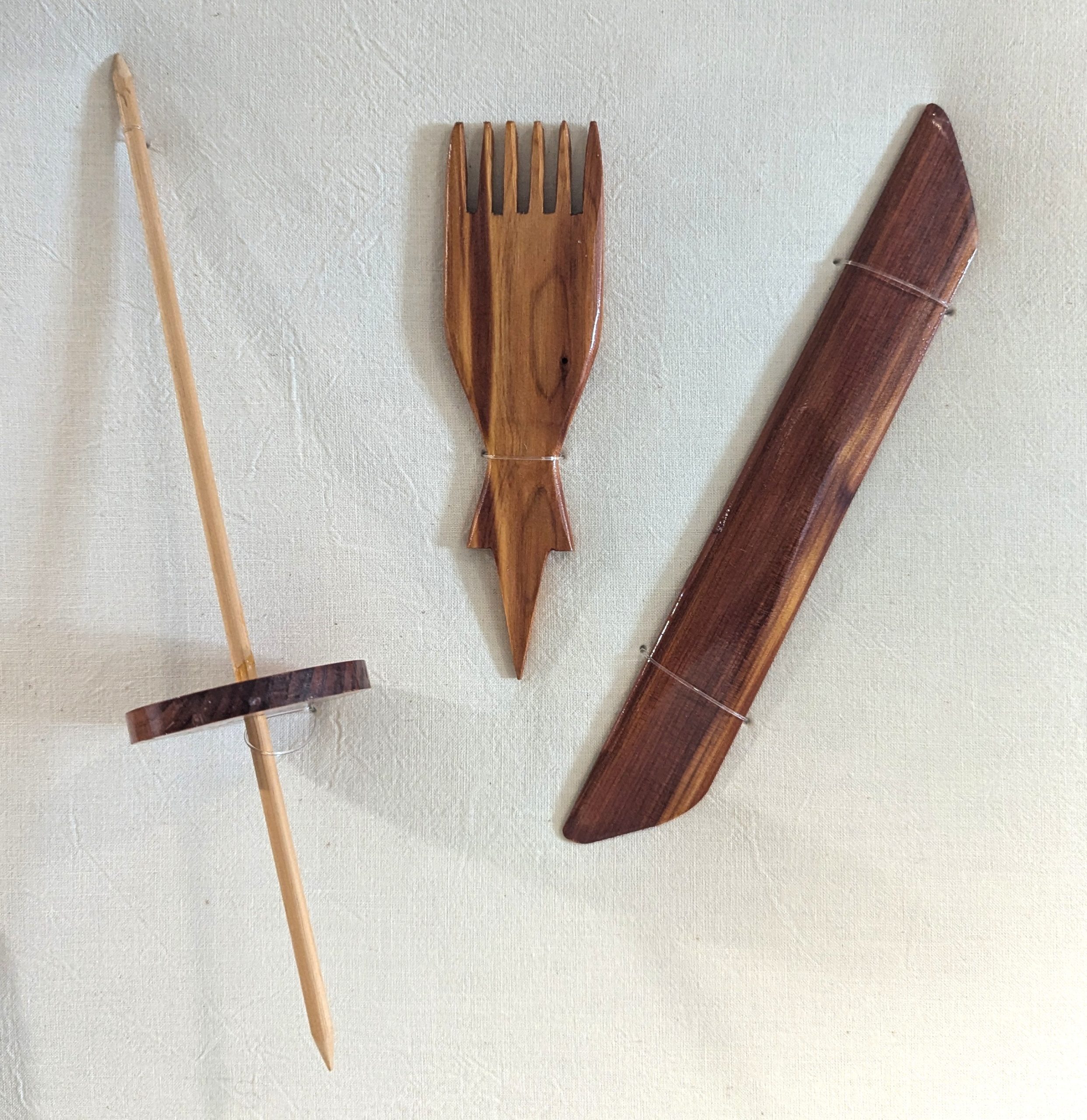 Three small wooden tools, including a drop spindle, a comb, and a simple batten used to straighten a row of weaving.