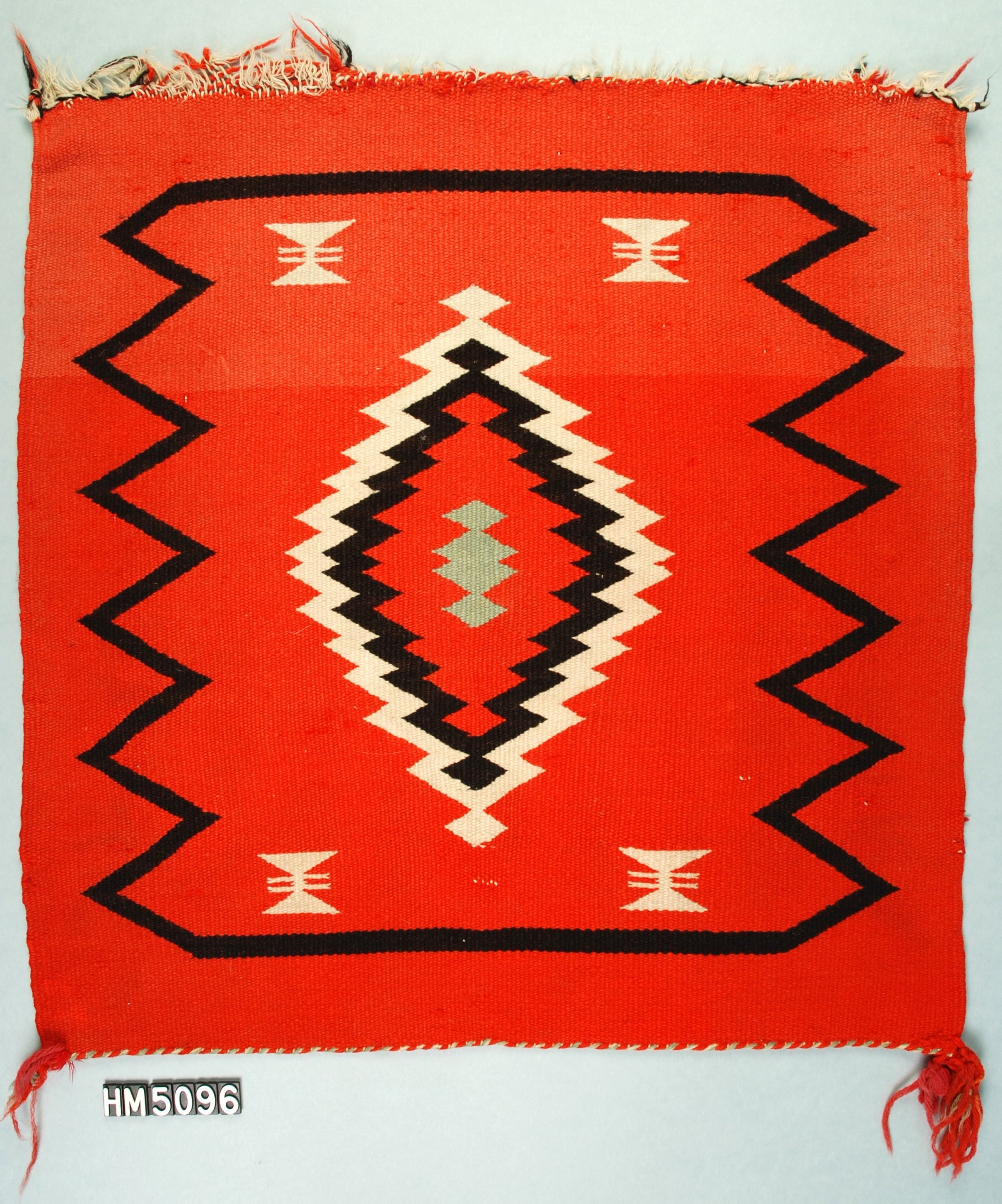 Small woven textile in red, black, and cream with a diamond-shaped central motif with serrated edges.