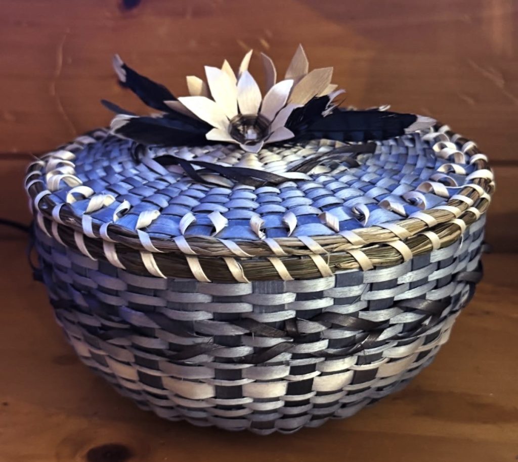 A basket with a cover. The basket is woven with purple and natural colors and includes intricate curls and patterns and a cluster of flowers at the center of the cover.