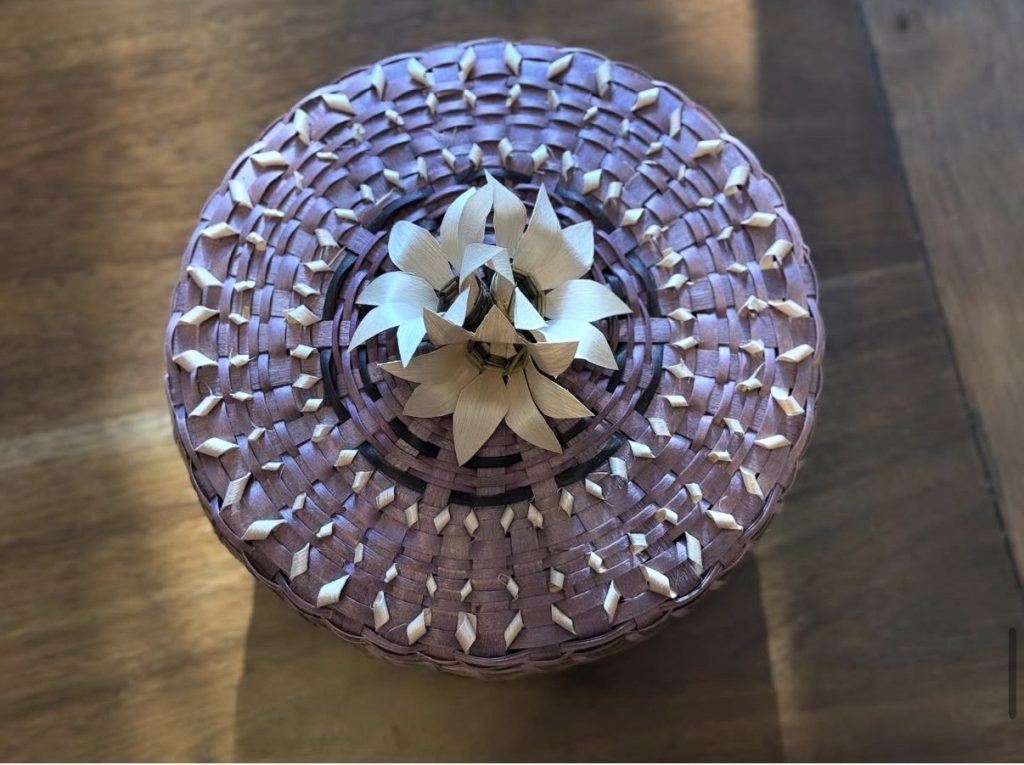 A top-down view of a basket woven in purple and natural colors with intricate curls and a cluster of flowers at the center of the cover.