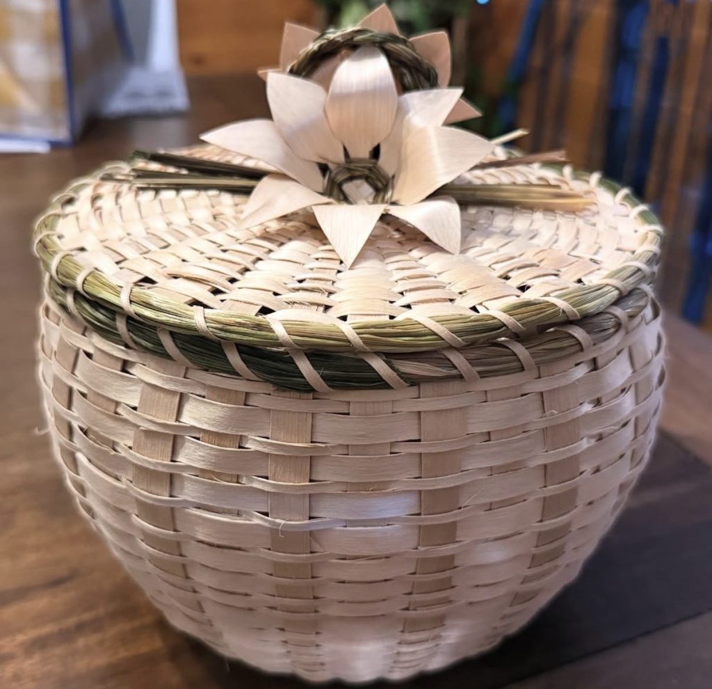 A basket woven in natural wood with grass lashed at the rim and a cluster of flowers with a loop of grass braid at the center of the cover.