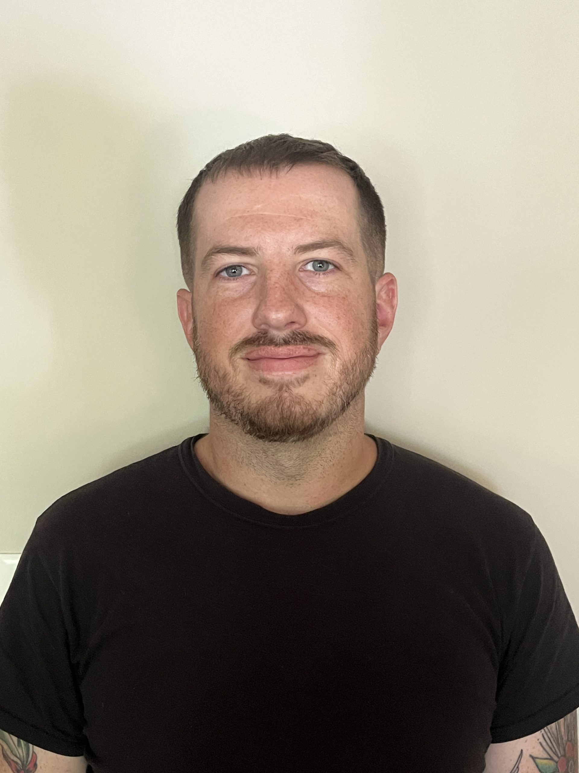 A Caucasian male with short hair and a short beard wearing a black shirt and smiling at camera.