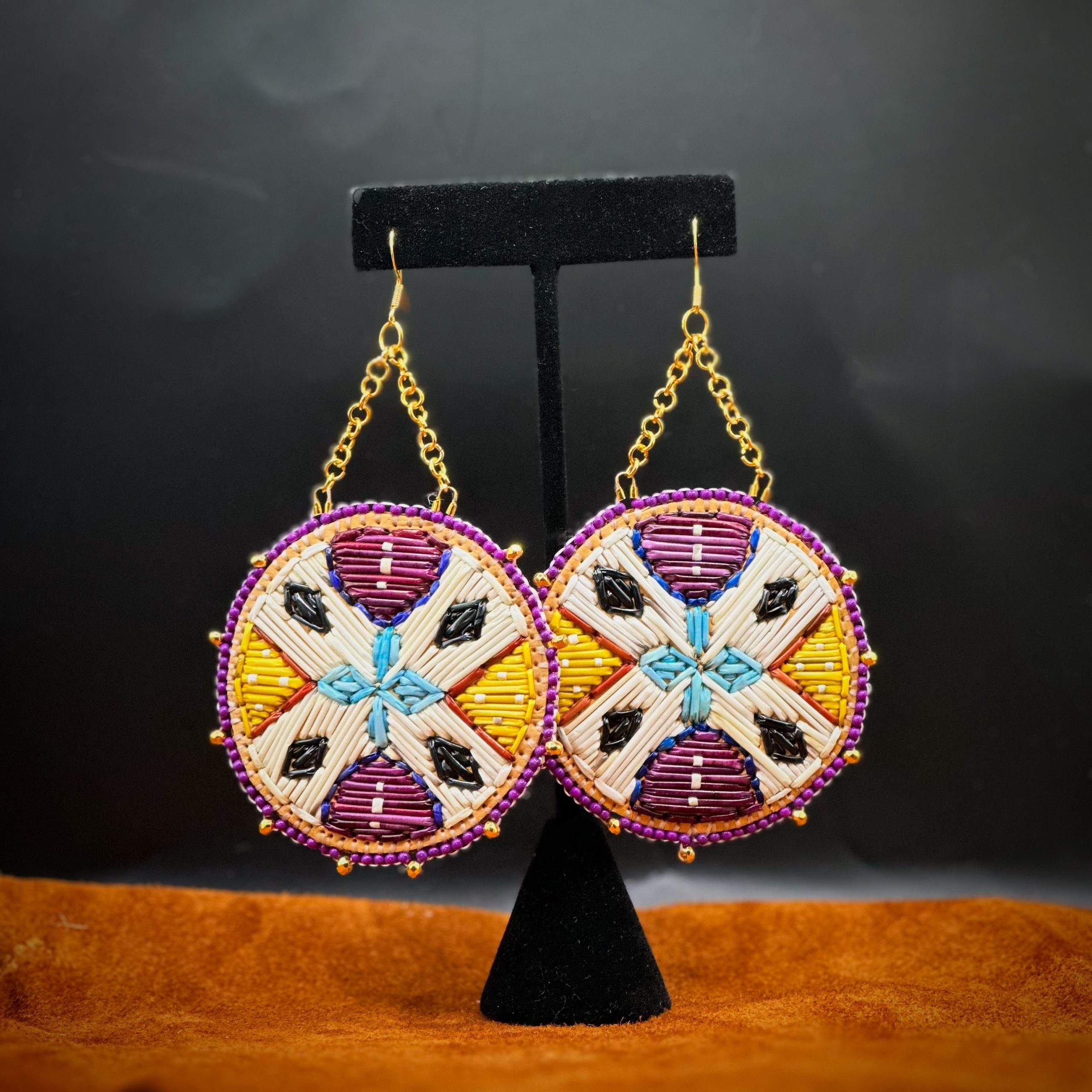 A pair of earrings hanging from a stand. They are each a circular and are decorated with quillwork in white, black, pale blue, purple, and yellow.
