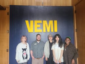 Four people smiling at camera underneath a sign that says "VEMI"