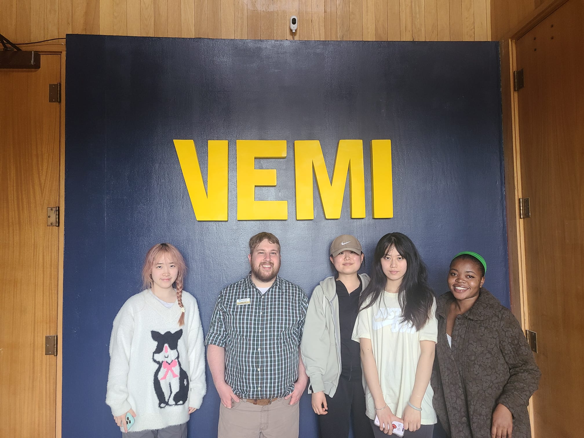 Four people smiling at camera underneath a sign that says "VEMI"