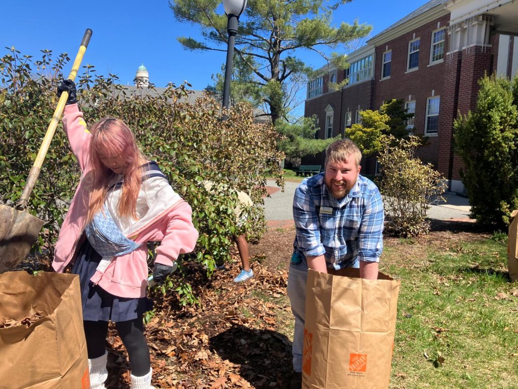 Student holding shovel to drop dead leaves into bag. Man standing over bag, smiling at camera while his hands are inside the bag, packing down leaves inside.