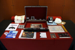 A table with a red tablecloth. Atop the cloth sits a suitcase with miniature versions of artwork laid out around and inside it.