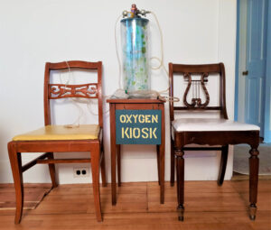 An 18” tall glass cylinder with a wooden lid. Attached to the lid is a small oxygen tank which hangs inside the glass. The valve apparatus that accesses oxygen is situated above the lid. There is an oxygen mask attached to one of the nozzles on the apparatus with plastic tubing. The entire assembly sits on a small wooden table bearing a sign on a wooden panel which says “Oxygen Kiosk” in yellow stenciled letters on a deep green background. On either side of the “Oxygen Kiosk” sits two wooden chairs.