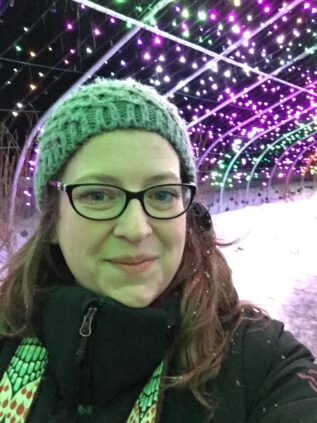 Jessica Brainerd stands in front of a set of colorful lights. She smiles a closed mouth smile. She wears glasses, a winter jacket, and a green knit cap.