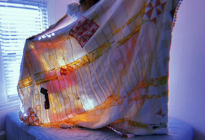 Artist stand holding white, pink and yellow quilt in front of her. Light streams through the window.