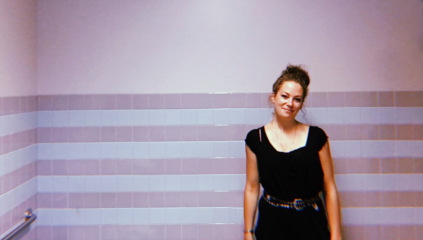 Kate stands in front of a pink topped, blue and purple striped wall. She wears black. Her brown hair is in a bun. She smiles closed mouth, with her head tilted.