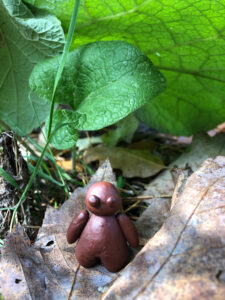 a small round clay figure is standing under a large green leaf looking out into the woods.