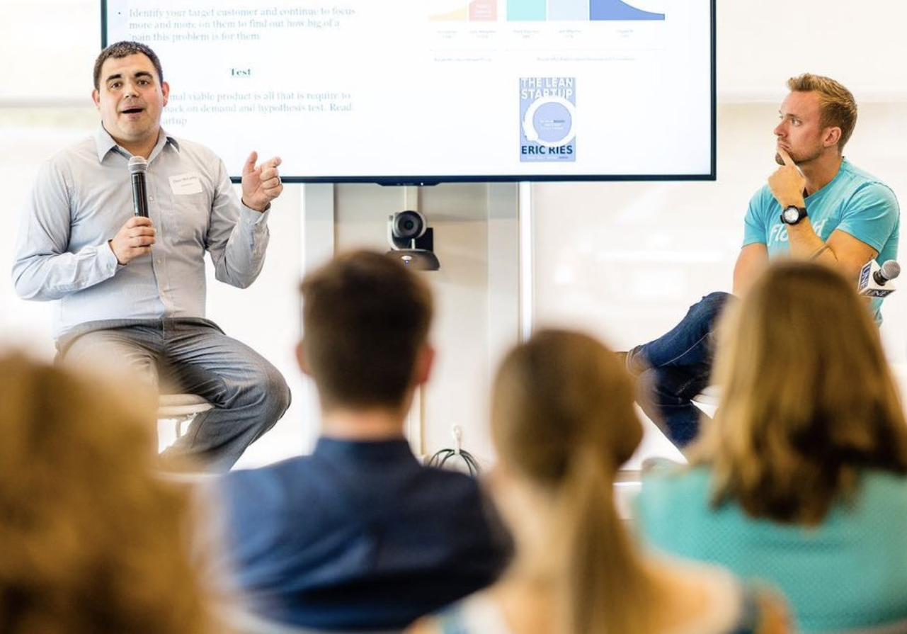Two young men sit on stools in front of a presentation screen