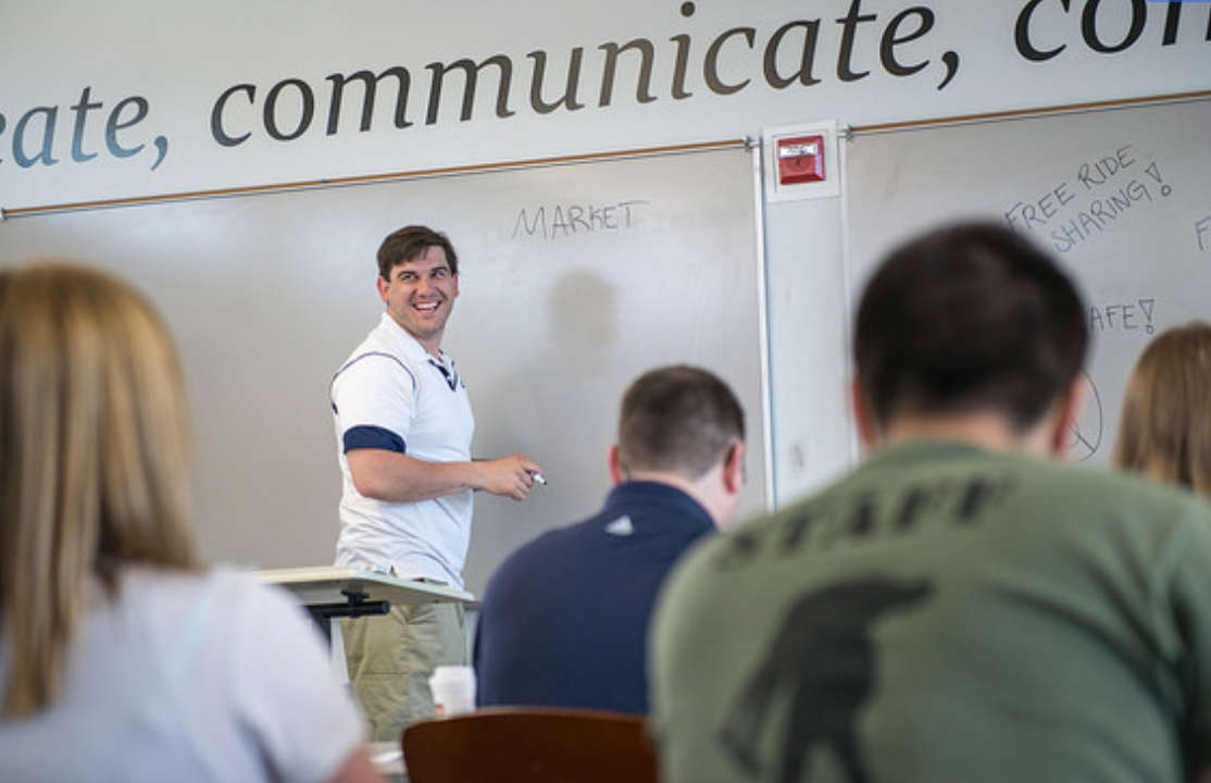 A smiling instructor at a whiteboard addresses a group of students at UMaine's Foster Center for Innovation.