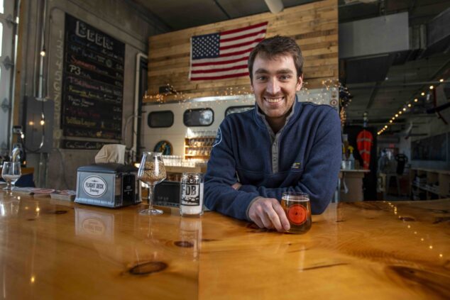 Nate Wildes standing behind the polished wood bar at Flight Deck Brewing in Brunswick, Maine.