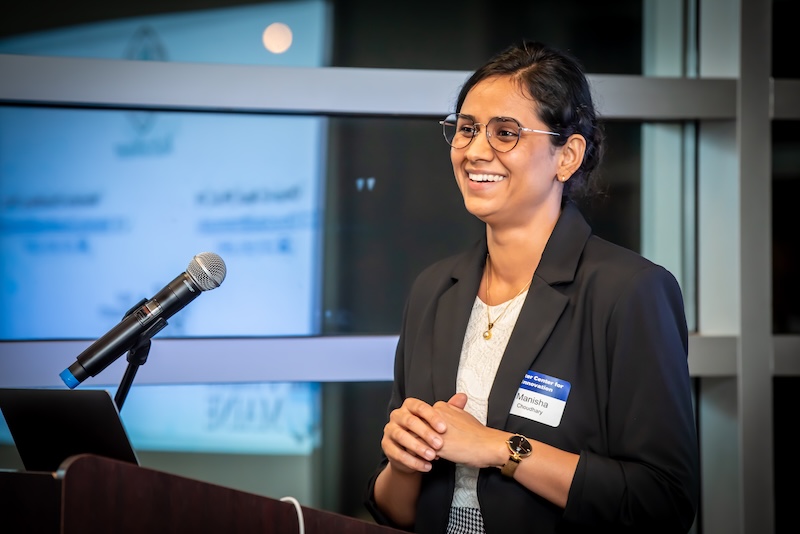 A woman in a black blazer and glasses smiles while standing behind a podium with a microphone.