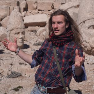 A man with long hair and a flannel shirt gestures as he stands in front of some ancient ruins.