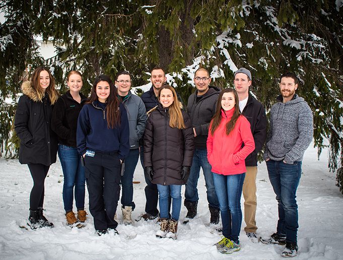Dr. Maginnis, 4 graduate students, and 5 undergraduates stand in front of snow-covered evergreens.