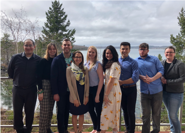 Dr. Maginnis and 8 lab members stand outside of MDIBL with the coast of Mount Desert Island behind them.
