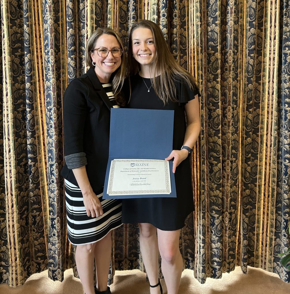 Dr. Maginnis and Avery Bond stand holding Bond's award for outstanding graduate research in molecular and biomedical sciences in 2024