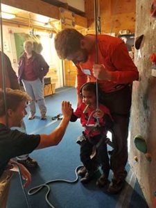 Young boy in indoor climbing harness giving high five to young adult