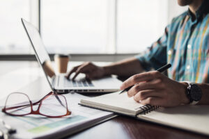 view of hand of professional in front of a laptop holding a pen ready to right in one hand and typing on laptop with the other