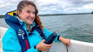 woman in blue life vest and light blue jacket records readings off side of a boat in the water on a cloudy day
