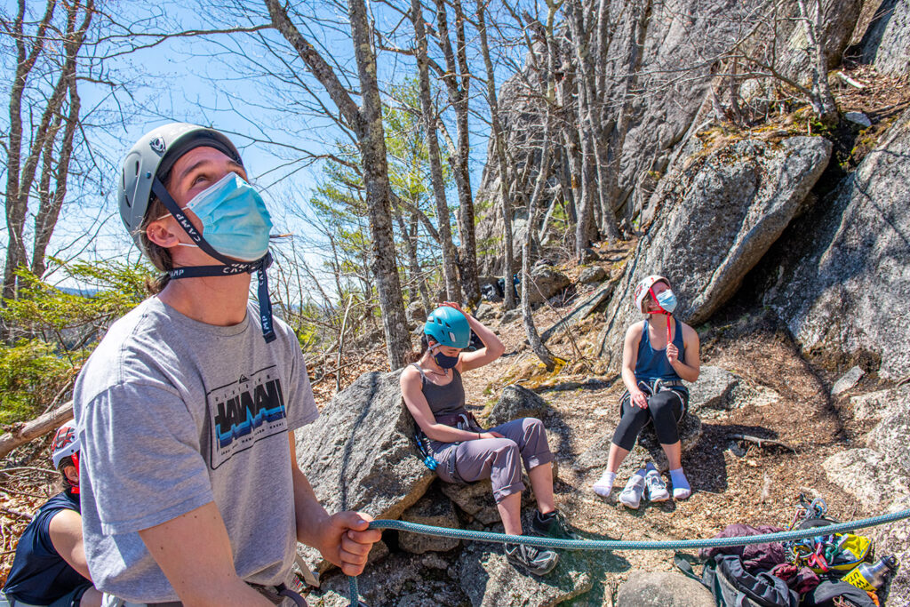 Students with rock climbing gear out in the Maine woods near a rock face.