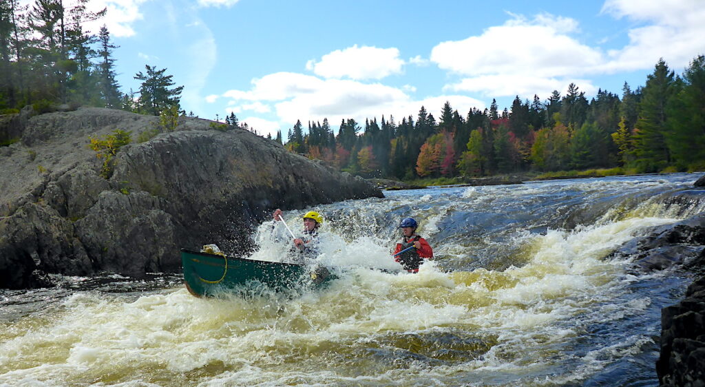 2 people in a green canoe paddle through whitewater under a blue sky fall day