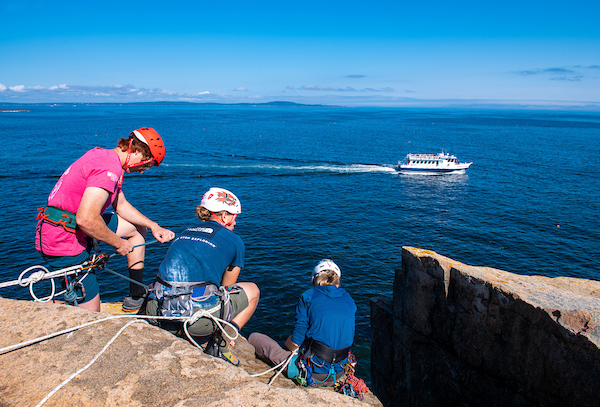 3 individuals in harnesses and helmets crouch on a cliff over the ocean