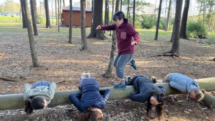 4 individuals lay on their stomachs on a telephone pole as another individual steps over them