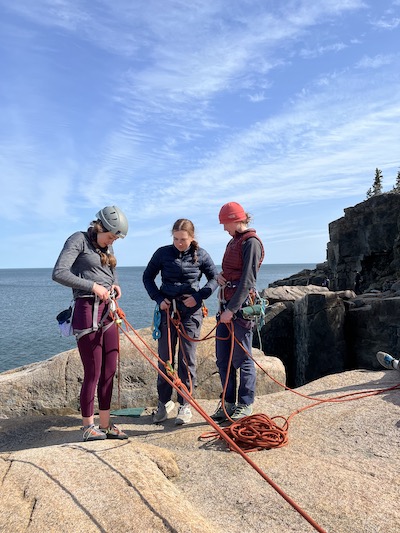 3 individuals in climbing harnesses stand on a cliff attached to ropes under a blue sky