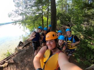 A group in yellow life jackets and helmets group together for a selfie in front of a body of water