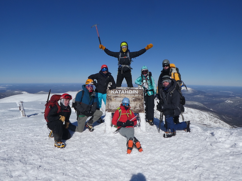 Winter group photo at the katahdin summit sign on a clear blue day.