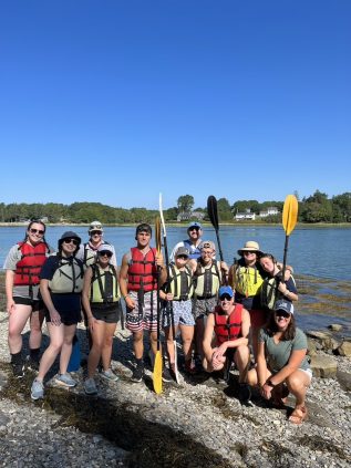 A group gathers together in front of the ocean holding sea kayaking paddles smiling