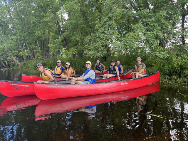 2 red canoes float under trees with students in them smiling for the camera