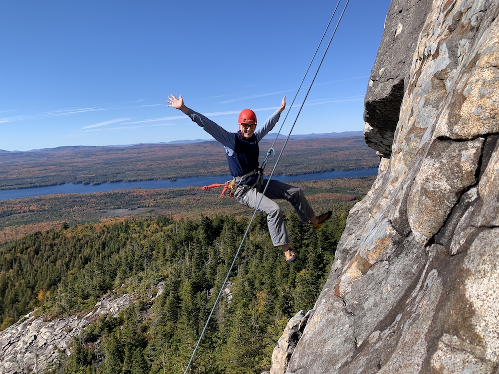 An individual hanging from a rope on a cliffside with arms out wide smiling