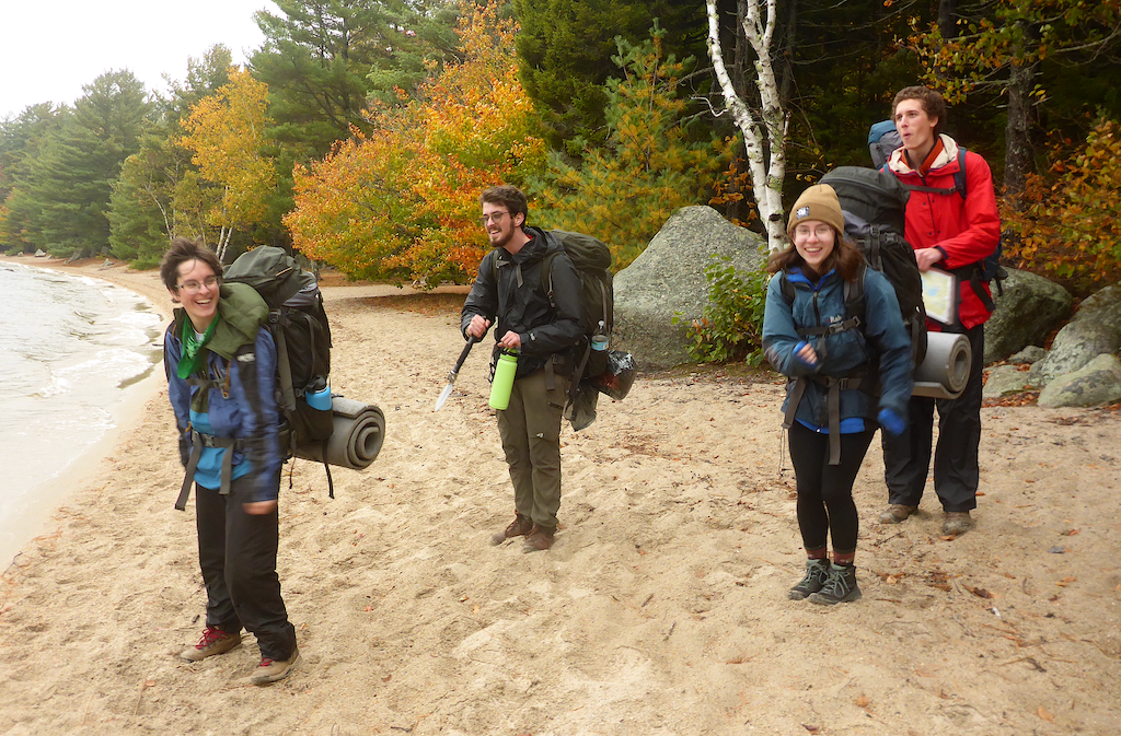 4 students standing on a lake beach with backpacks in the fall