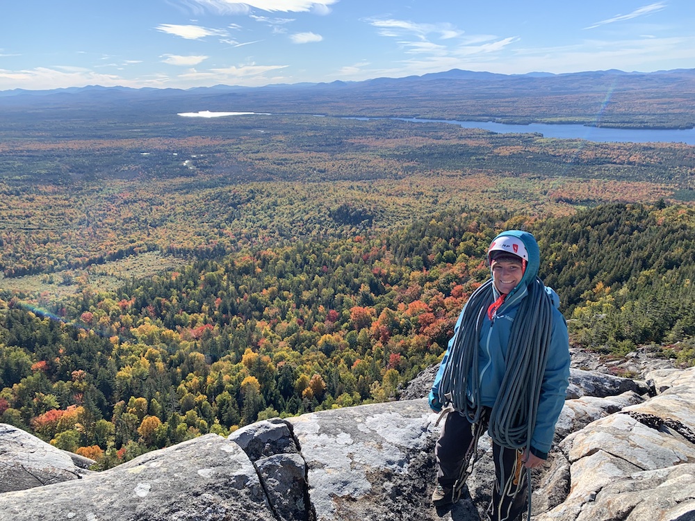 Person wearing blue jacket with hood up has climbing rope draped over her standing in front of fall colored trees