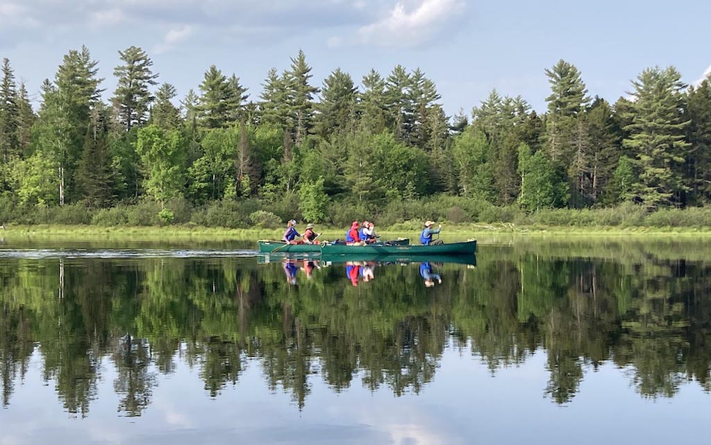 2 green canoes with people in them paddling down a still river lined in green trees