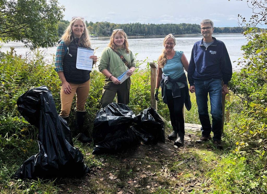 four smiling individuals standing on a grassy nook with the water in the background, with several bags of collected marine debris