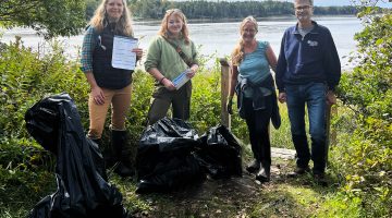 four smiling individuals standing on a grassy nook with the water in the background, with several bags of collected marine debris